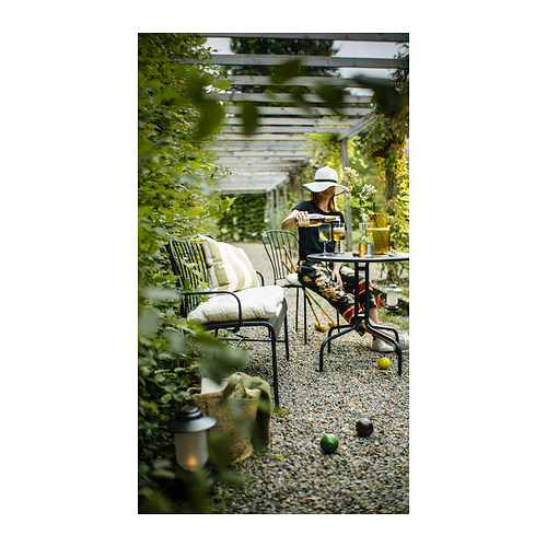 Mujer en el jardín del patio de diamantes, con sillas de metal negro LÄCKÖ, plantas en macetas, mesa con cítricos, vasos y velas.