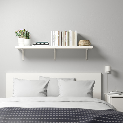 Dormitorio minimalista con estantería BERGSHULT que contiene libros, plantas, cestas. Cama blanca, cabecero gris, lámpara de noche.