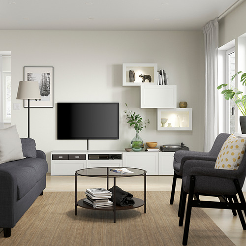 Sala de estar moderna con mueble de TV SINDVIK blanco, sofá azul, silla gris y plantas. Mesa de centro de cristal, grandes ventanales.
