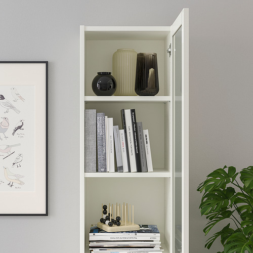 Una estantería blanca BILLY con puertas de vidrio exhibe libros y jarrones. Su diseño permite un almacenamiento sin polvo y un uso versátil.