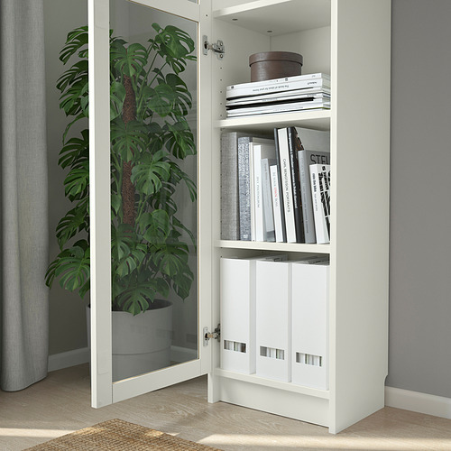 Una estantería BILLY con puertas de vidrio para libros y los mantiene libres de polvo. A su lado se coloca una gran planta de interior.