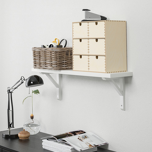 Un estante de pared blanco contiene una caja de madera, una cesta con tijeras y una calculadora. Abajo, un escritorio con una lámpara, un jarrón y revistas.