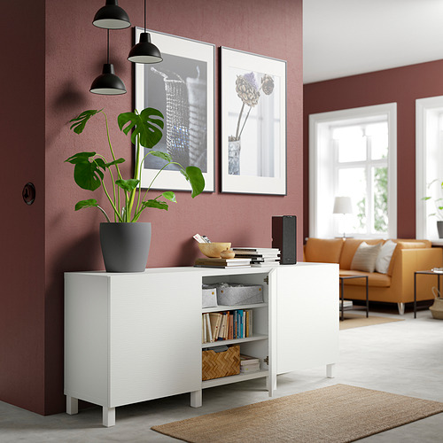 Mueble de almacenamiento blanco BESTÅ con estantes ajustables, con libros y una planta, frente a una pared roja con obras de arte enmarcadas.