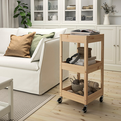 Carro de almacenamiento de madera junto al sofá, que contiene artículos de tejido, libros y canasta. El pino claro complementa el sofá blanco y el suelo de madera.