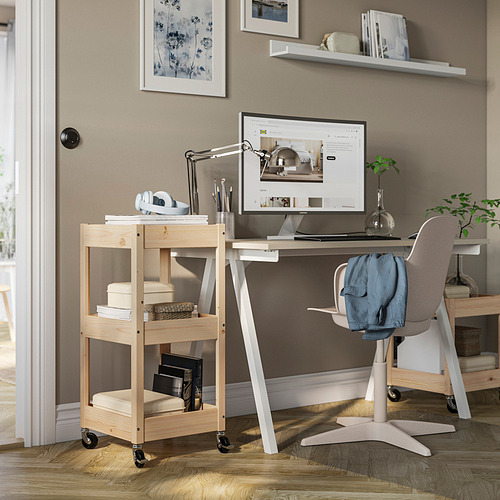 Oficina en casa con carrito de madera, escritorio y monitor. Paredes ligeras, carro de pino con estantes. Silla gris con chaqueta.