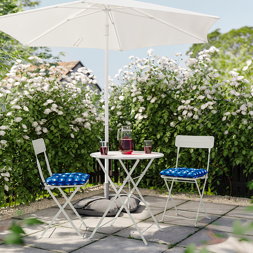 Pequeña instalación al aire libre con mesa y sillas plegables blancas, cojines azules, flores blancas y una jarra de bebida bajo una sombrilla blanca.