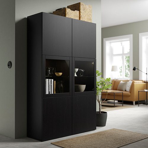 Mueble de almacenaje BESTÅ negro con puertas de cristal, donde se guardan objetos de decoración y libros. Se alza junto a una sala de estar con un sofá, una lámpara y una planta.