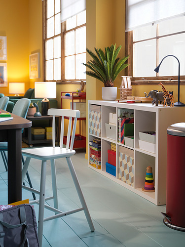 Habitación luminosa con una pared amarilla, una silla junior blanca, una estantería de juguetes y una mesa de madera. Los grandes ventanales dejan entrar la luz natural.