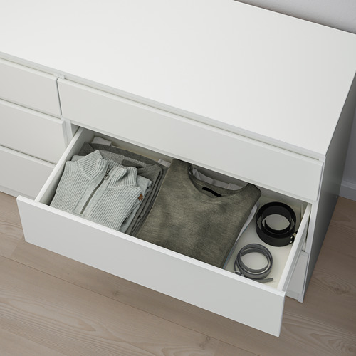 Cajón de la cómoda blanco abierto que revela ropa y artículos de cocina cuidadosamente doblados.