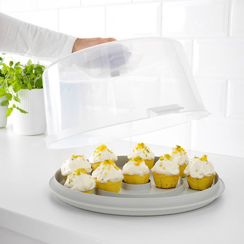 La mano sostiene la tapa transparente del portador de pasteles KRISPIG, mostrando cupcakes de limón con glaseado en la encimera blanca.