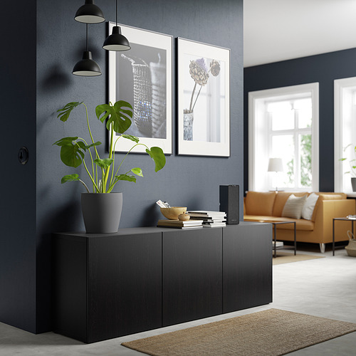 Salón moderno con aparador BESTÅ negro, libros, plantas, obras de arte, sofá marrón, lámparas colgantes.