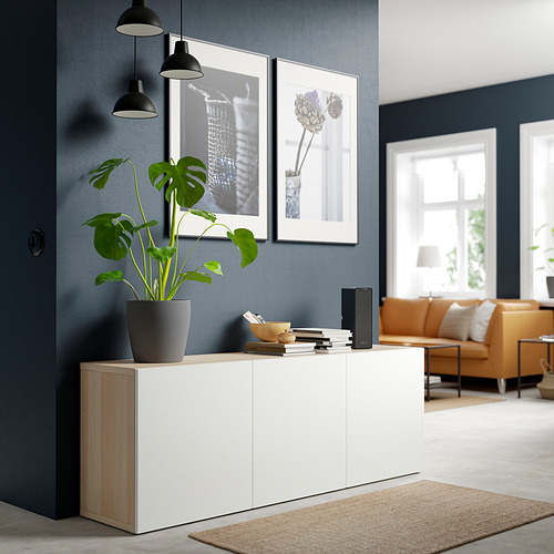 Sala de estar moderna con aparador BESTÅ, planta, sofá naranja, alfombra y cuadros en una pared azul oscuro.