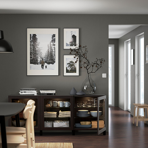 Comedor moderno con paredes oscuras, un aparador negro con platos, tazas y jarrones decorativos. Arte montado en la pared y luz natural desde el pasillo.
