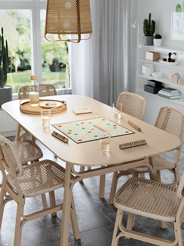 Moderno juego de comedor con tablero de scrabble, maderas claras, sillas de mimbre, luz natural.