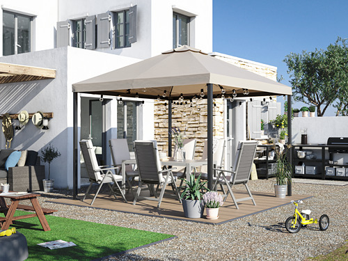 Patio trasero con tarima RUNNEN beige, sillas grises, mesa y toldo con luces, rodeado de grava y vegetación.