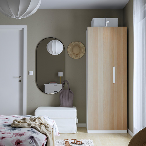 Minimalist bedroom with wooden PAX wardrobe, white dresser, and large mirror. Floral quilt and lamp add color. Wardrobe offers efficient storage.