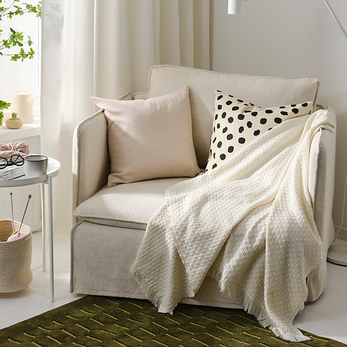 Manta HORNMAL beige en el sillón, almohadas, mesa auxiliar con taza, tejido, planta, alfombra verde - ambiente acogedor.