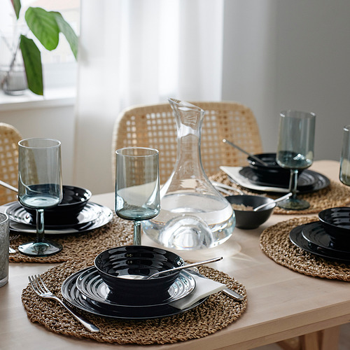 Mesa con vajilla de cristal negro, apilable y apta para lavavajillas, sobre manteles individuales tejidos con vasos transparentes y jarra de agua.