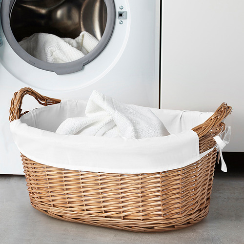 La cesta de lavandería TOLKNING en tejido de sauce natural con una gran bolsa interior blanca. De forma circular, con asas resistentes, cabe al lado de una lavadora.