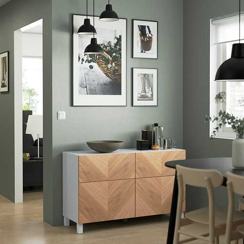 Sala de estar moderna con aparador de madera clara BESTÅ, fotos y vegetación. Incluye cuenco de piedra, jarras, mesa de comedor y sillas de madera.