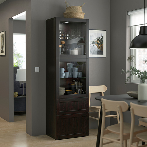 Moderno gabinete de almacenamiento de madera oscura y vidrio, más alto que las sillas cercanas, exhibe tazas y vasos, con canasta tejida en la parte superior.