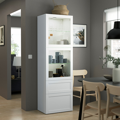 Moderno armario blanco BESTÅ con puertas de cristal, que almacena tazas y cuencos azules. Alto, rectangular, de madera y cristal, encaja bien en el comedor.