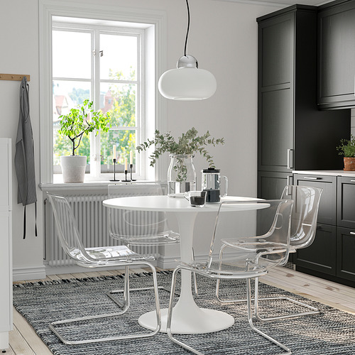 Pequeño y moderno juego de comedor con mesa redonda blanca y sillas transparentes. Cocina adyacente, iluminación brillante.