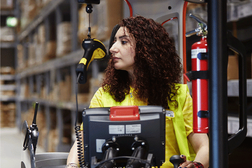 A female warehouse worker with curly hair wearing a bright yellow safety vest operates a forklift vehicle inside a self-serve warehouse.