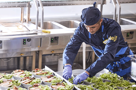 An IKEA food service employee wearing a blue uniform, hat, and gloves carefully prepares several chicken salads in a commercial kitchen cafeteria line.