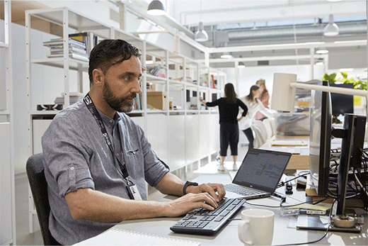 A man with a beard wearing an IKEA lanyard sits at a desk typing on a keyboard in a bright, modern office setting.