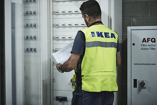 A view from behind of an IKEA maintenance worker wearing a neon yellow safety vest, holding technical documents while inspecting a large electrical panel.