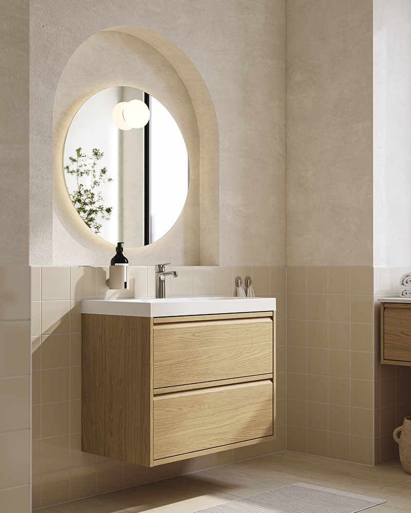 An oak eect, two-drawer ÄNGSJÖN/BACKSJÖN washstand is on a wall below a round mirror in a modern beige and white bathroom.