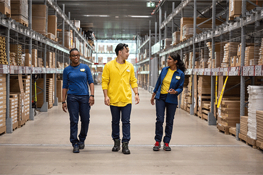 Three IKEA co-workers wearing blue and yellow uniforms walk and chat together down a wide aisle of a self-serve furniture warehouse.