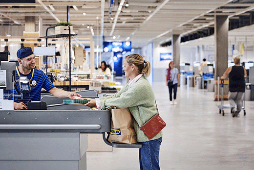 A smiling IKEA cashier in a blue striped uniform assists a female customer who is packing items into a paper bag at a store checkout lane.