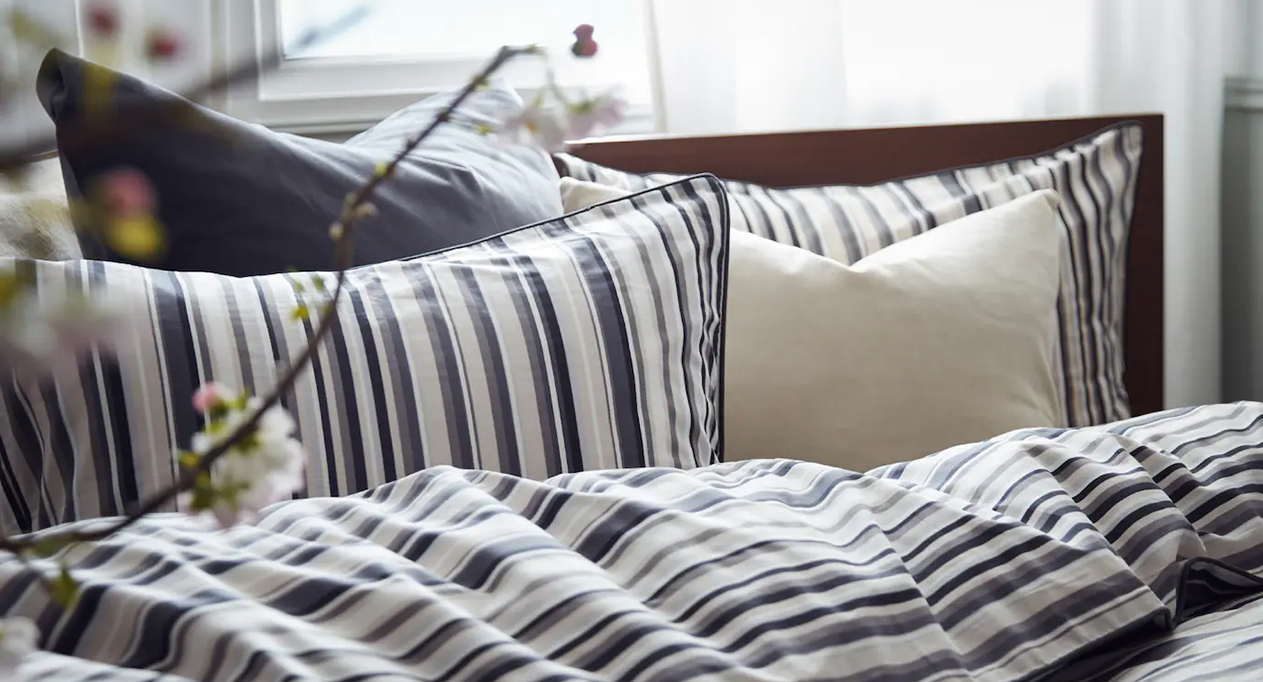 Cama acogedora con ropa de rayas, almohadas suaves y flores cerca de una ventana iluminada por el sol.