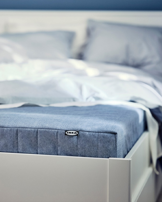 Corner of a white modern style bed, with the mattress visible in blue and a white quilt on top.