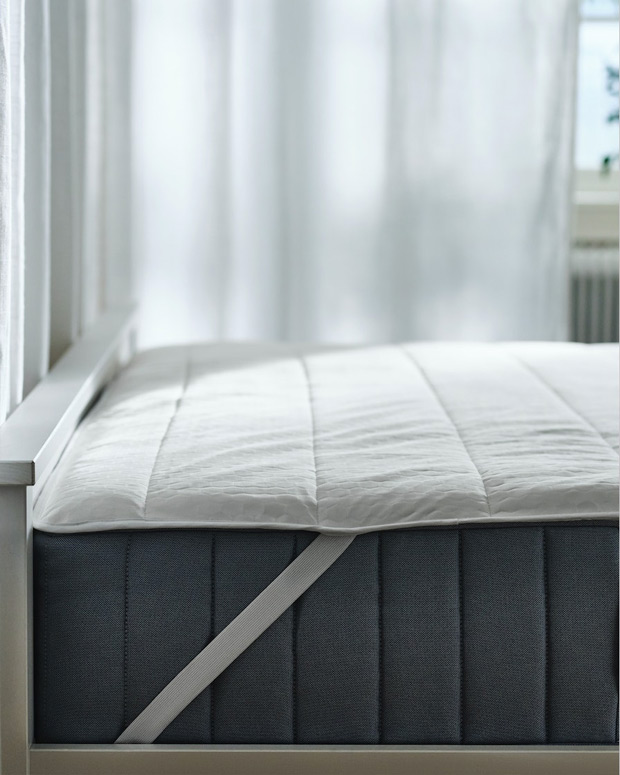 Image of a bed with a gray mattress visible, and the mattress protector with the tape holding it in place.