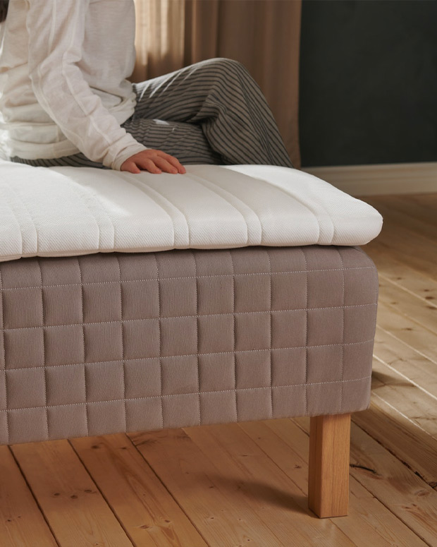 a person sitting on a bed, the mattress is visible, it looks big and on top of it there is a small mattress, the image only shows the corner of the bed and only part of the body of the seated person.