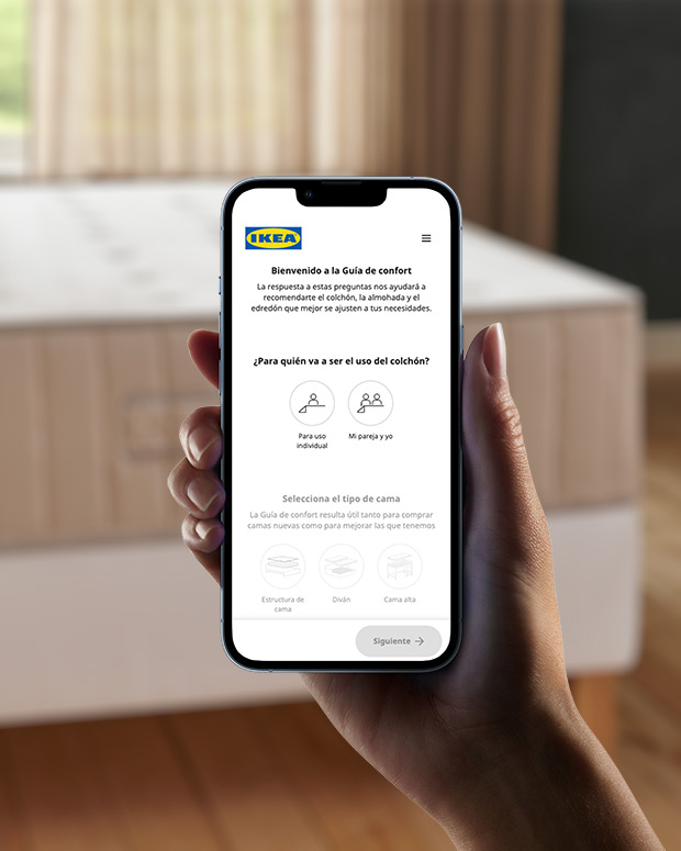 Hand holding a cell phone, on the screen you can see a mattress guide app, in the background out of focus you can see a large beige mattress.