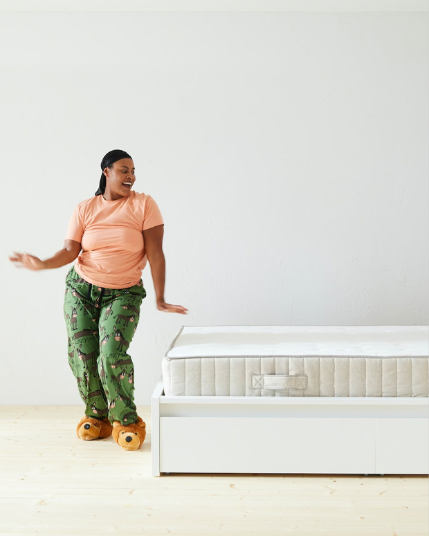 Woman standing in a dynamic pose in front of a bed with a white mattress visible, the shot is fully illuminated and white.