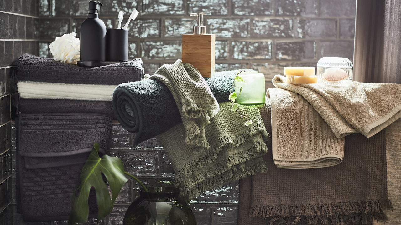 A bathroom with green tones featuring several sets of white and black VÅGSJÖN towels, along with a GANSJÖN bathroom set and a DRAGAN soap dispenser.