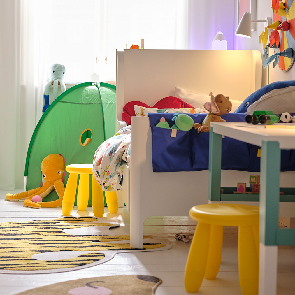 A children's room with a TROFAST storage system on the left, a PAHL desk on the right with two yellow MAMMUT stools, and a DVÄRGMÅS tent in the background.
