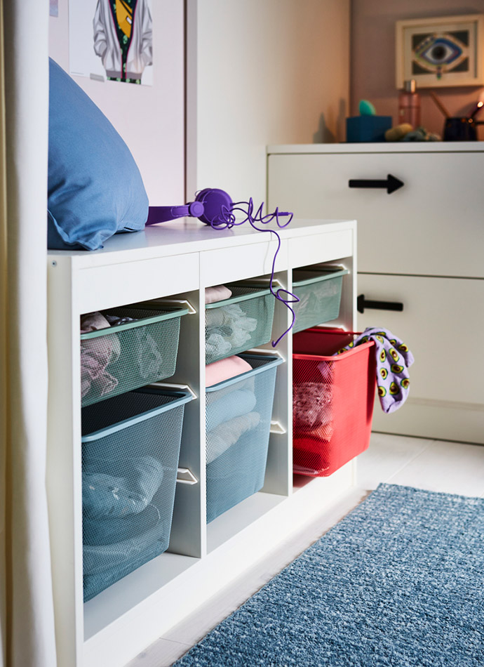 The image shows a white furniture unit with blue and red mesh baskets, purple headphones, and a blue rug, creating an organized atmosphere.