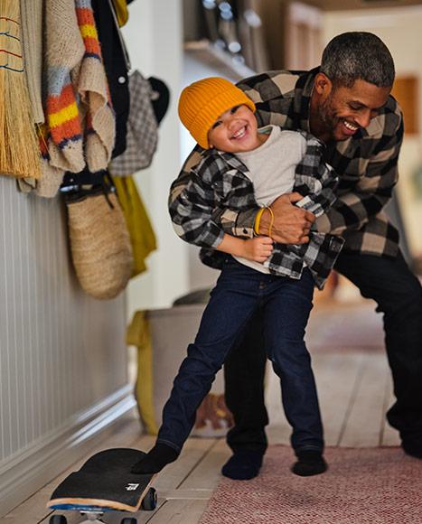 A smiling man helps a child with a yellow beanie balance on a skateboard in a hallway.