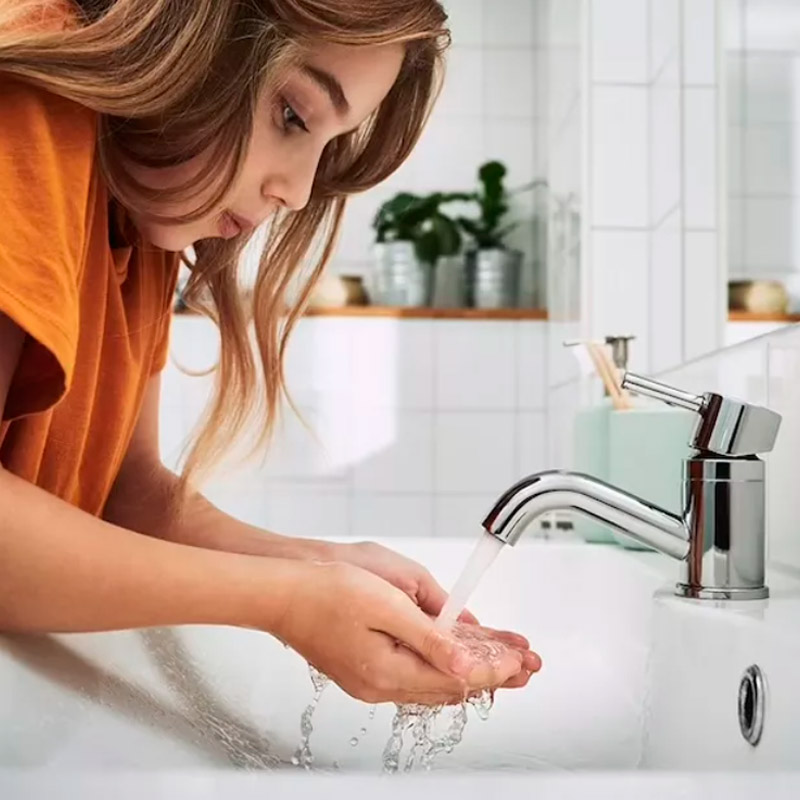 Young girl washing hands at a modern sink with running water in a bright, tiled bathroom with plants in the background.