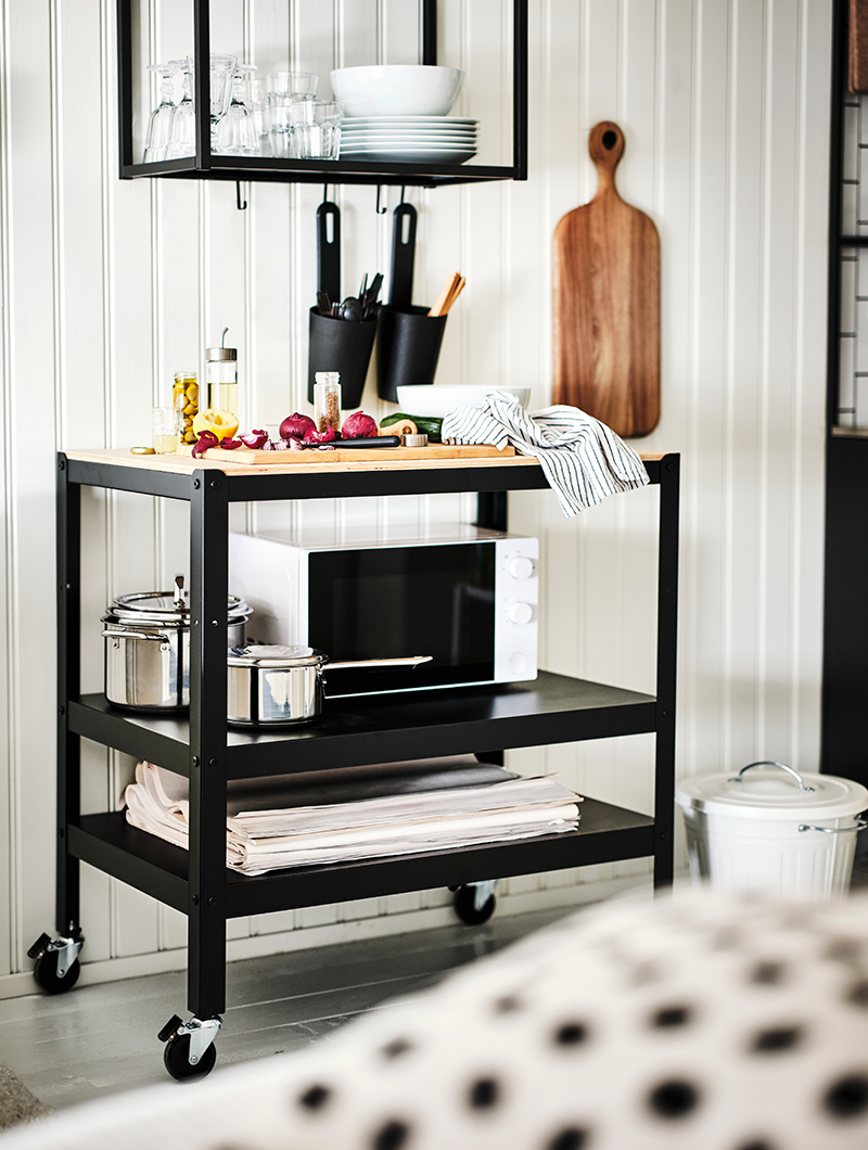 Black kitchen cart with pine worktop, underneath a small oven and several pans and on top a cutting board with food to prepare.