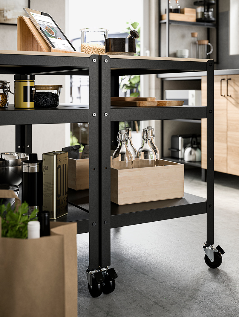 High kitchen cart, underneath it has wooden boxes with bottles inside, on top there is a graphic tablet with a cutting board and jars with lids.