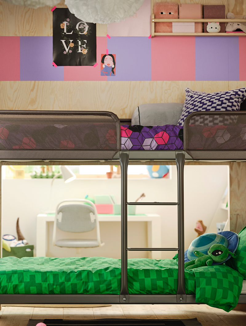 Metal bunk bed in a youth bedroom, with green and purple bedding. The light wood wall features a pink and lilac pattern.
