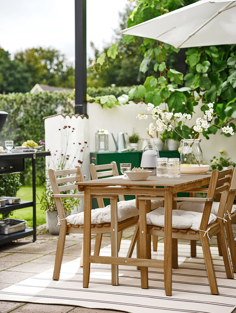 Outdoor dining room under an umbrella, on top of it you can see jars with lids, crockery and vases, in the background you can see a barbecue.