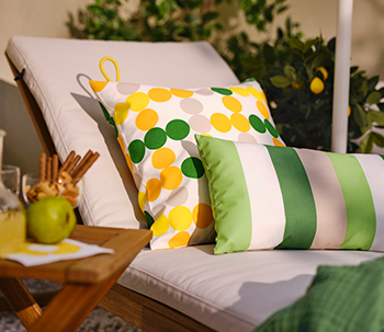 Tumbona con cojines blancos y decorada con dos cojines de colores: uno con lunares amarillos y verdes, y otro con rayas verdes, junto a una mesa con bebida y frutas.